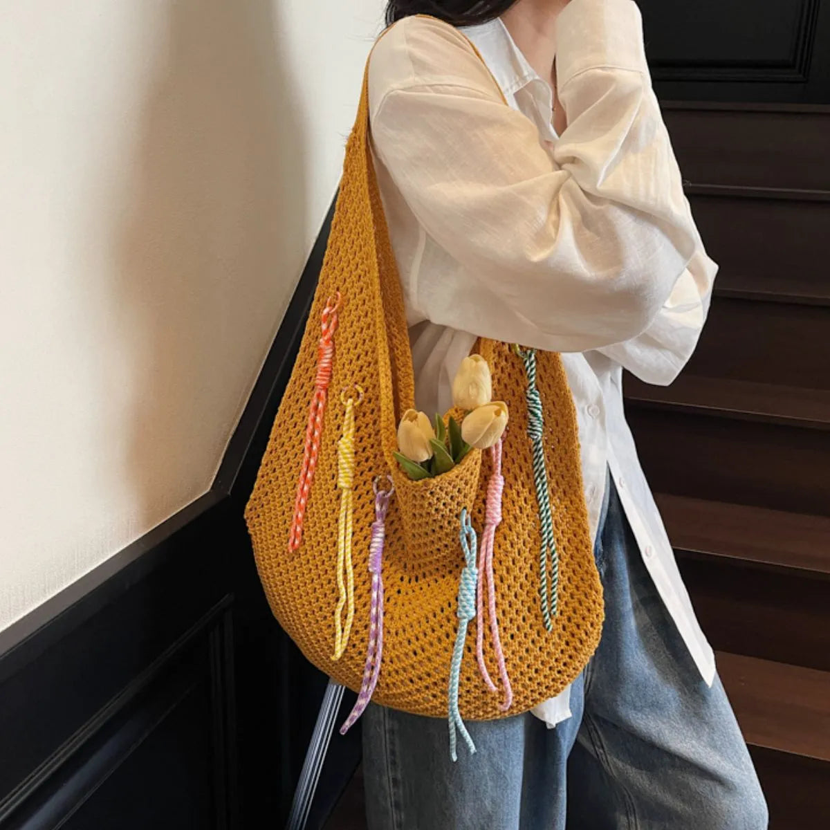 Person holding a yellow crochet bag with colorful strings and tulips, standing indoors.