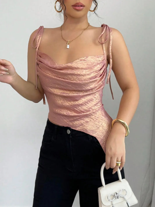 Woman wearing a pink sparkly top and black pants, holding a white handbag against a gray background.