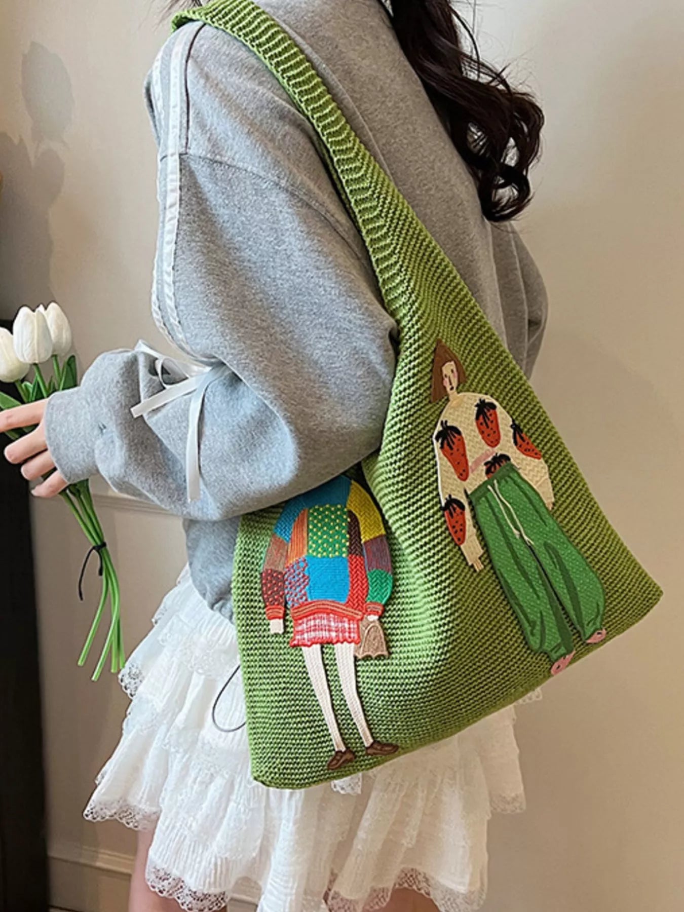Person holding a green bag with artistic designs, wearing a gray sweater and white skirt.