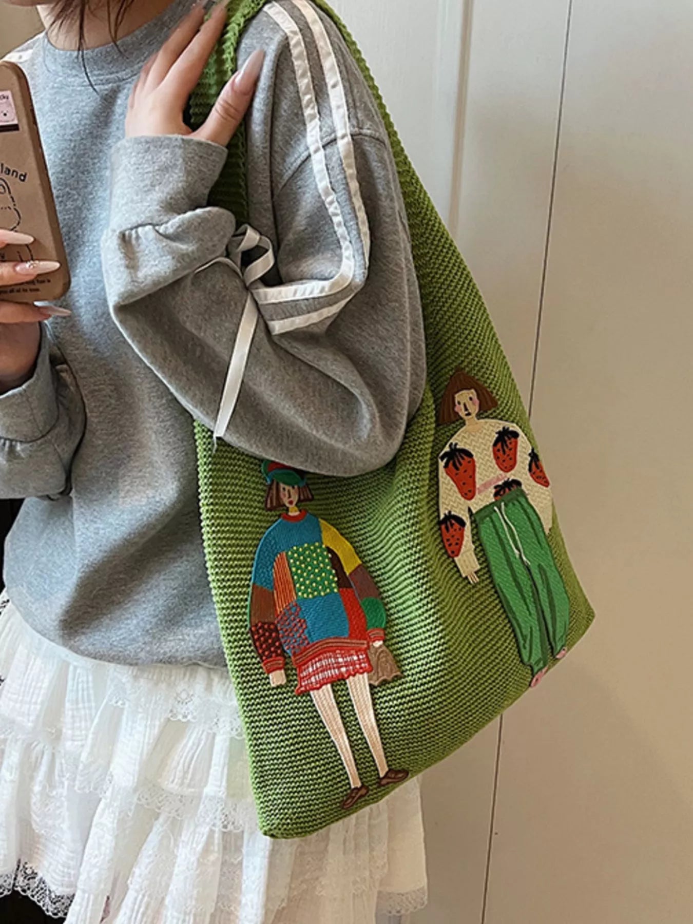 Green knitted bag with colorful appliqués held by a person wearing a gray sweater.