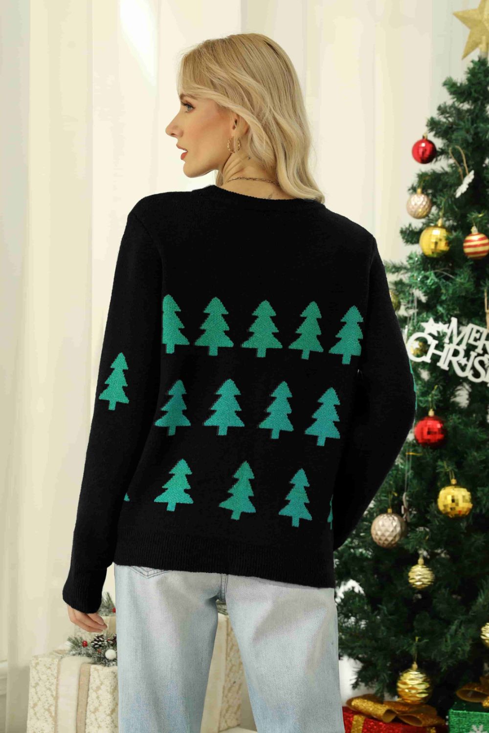 Person wearing a black sweater with green Christmas tree patterns in front of a decorated Christmas tree.