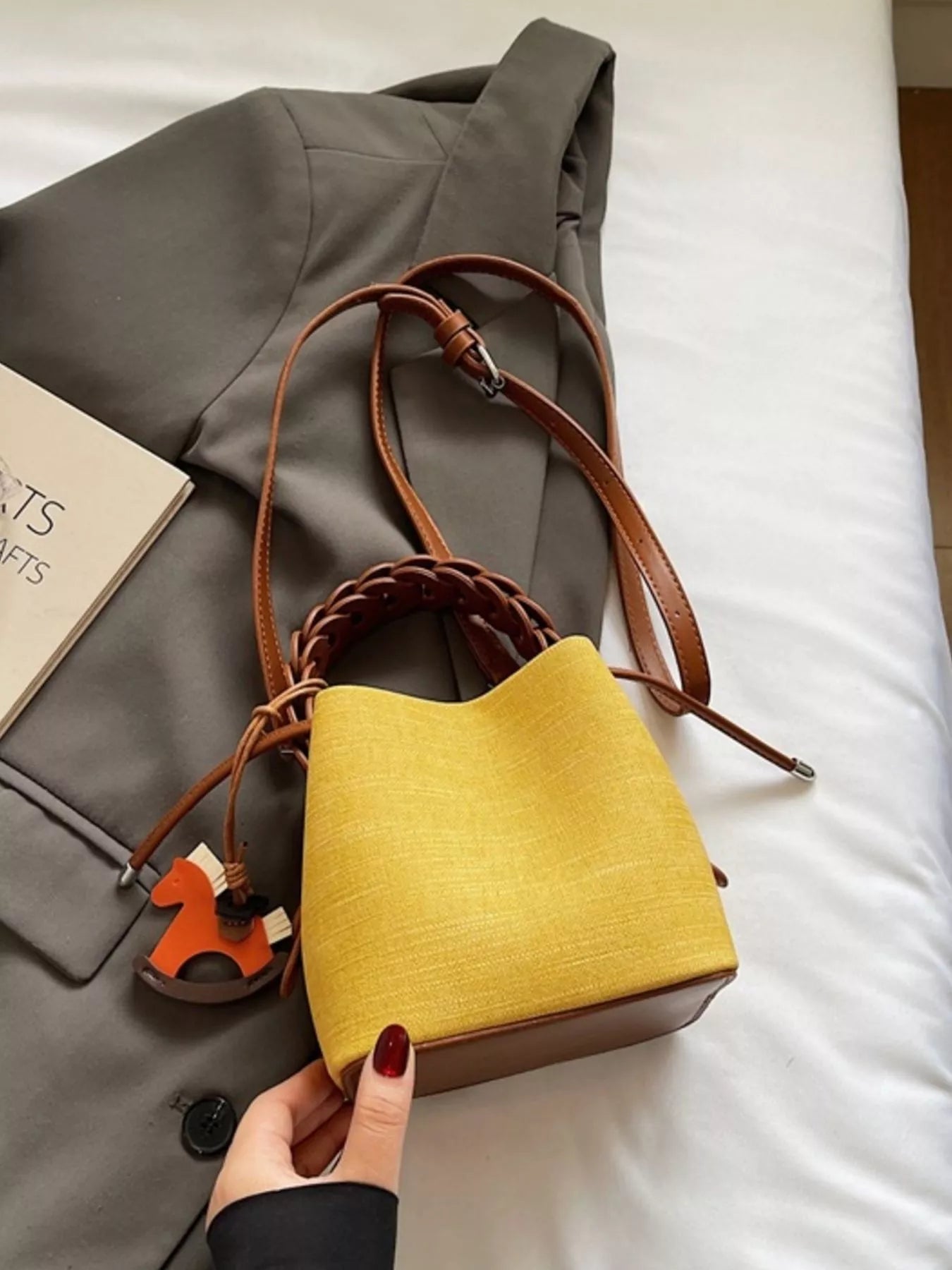 Yellow handbag with brown handle held by a hand on a white surface next to a gray coat.