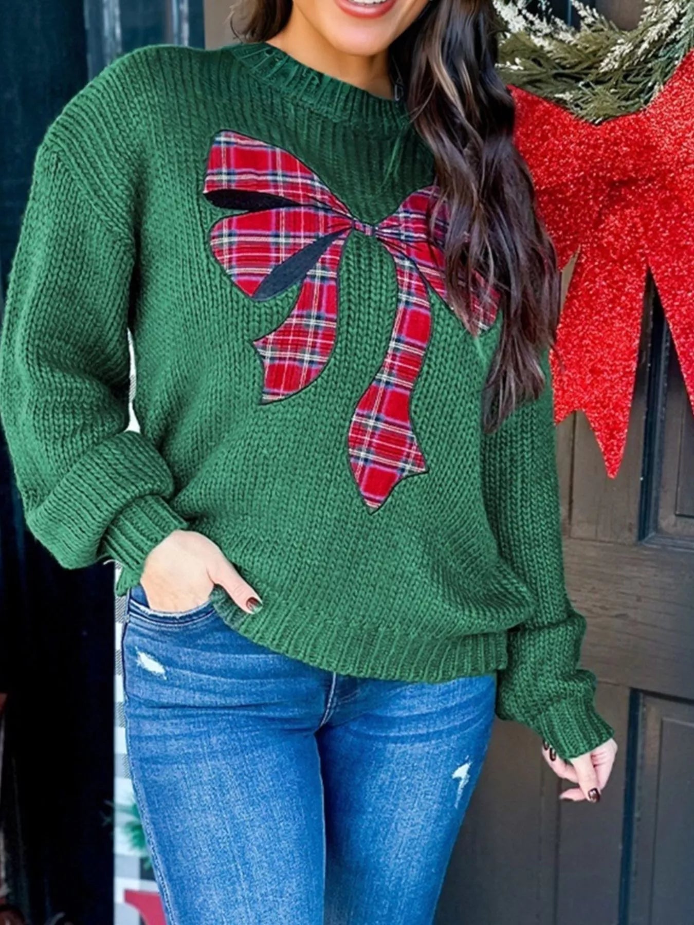Person wearing a green sweater with a large plaid bow design, standing next to a Christmas tree.