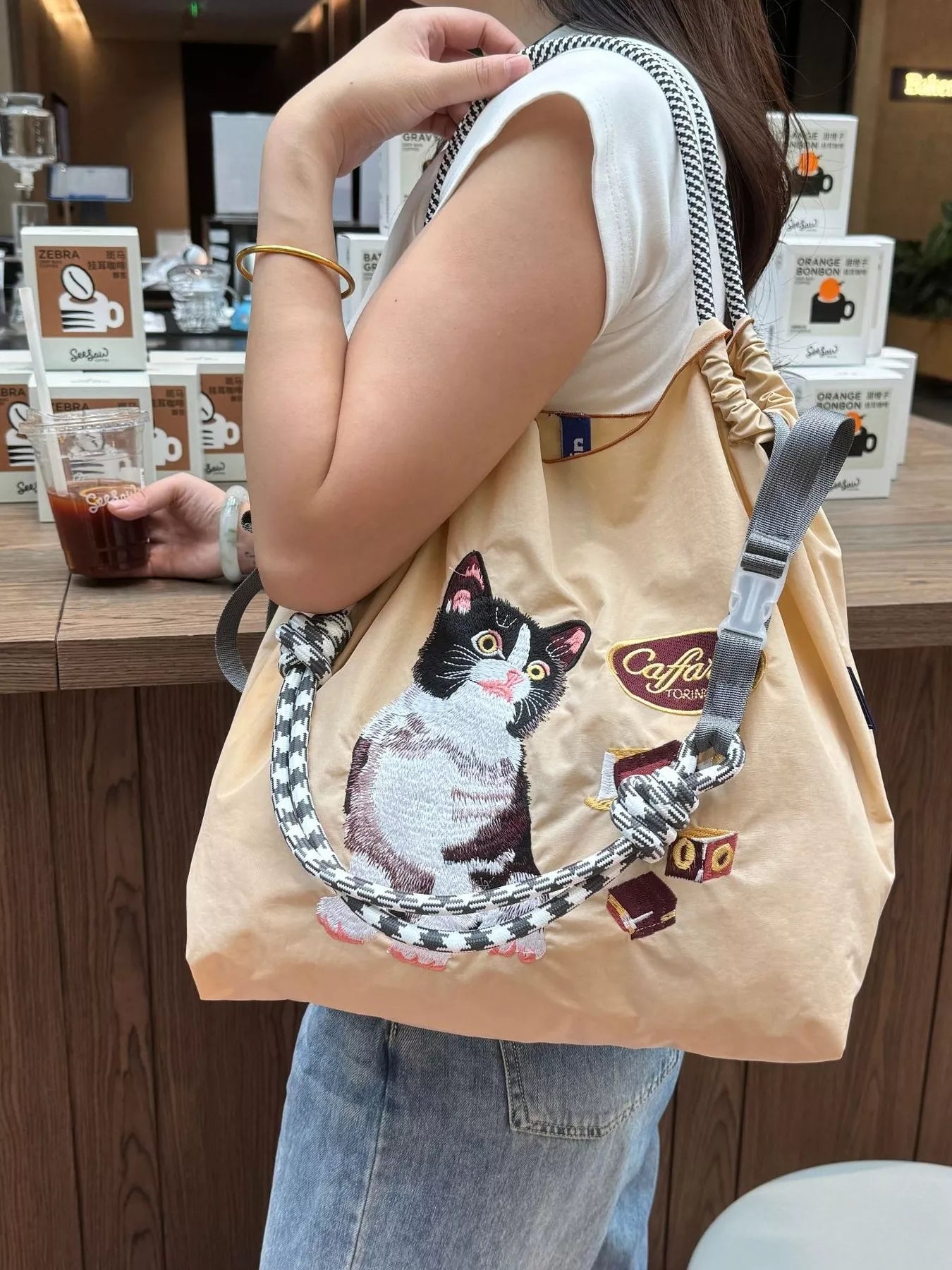 Person holding a beige tote bag with a cat design in a cafe setting